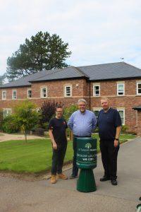 Three men standing with a giant version of St Teresa's Hospice's green collection tins. The photo is taken in the picturesque hospice gardens.