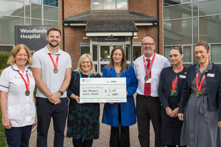 Group of seven people, five of whom are wearing Great North Run medals, stood outside Woodlands Hospital in Darlington. They are holding a cheque for three thousand and ninety pounds written out to St Teresa's Hospice.