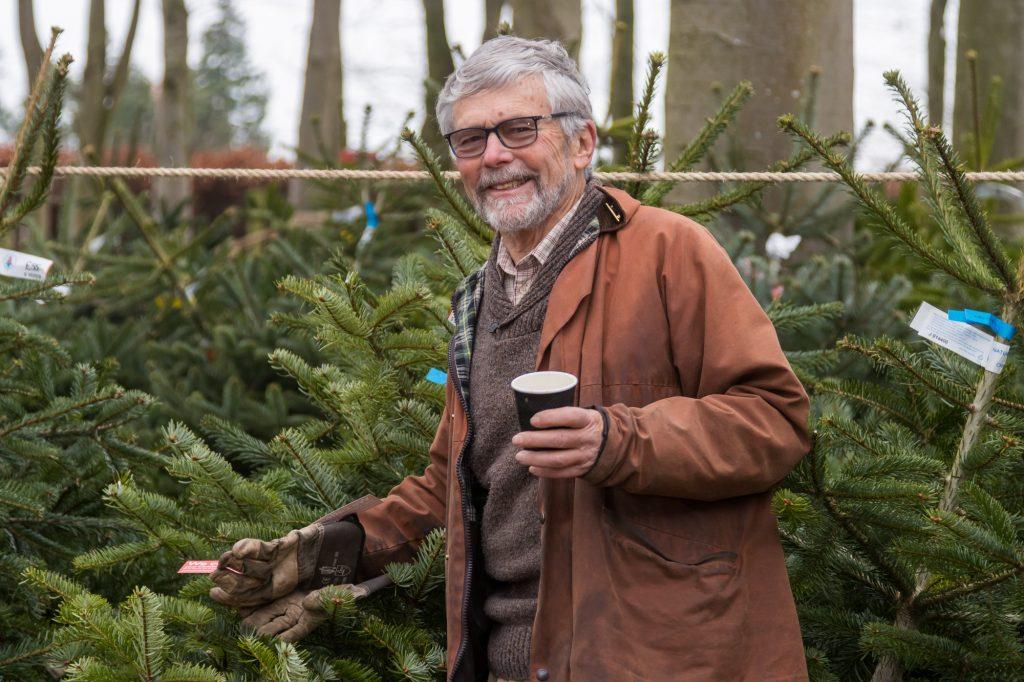 Close up of a man smiling at the camera holding a cup of coffee in one hand and a Christmas tree in the other. There are Christmas trees in the background.