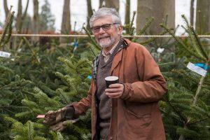 Close up of a man smiling at the camera holding a cup of coffee in one hand and a Christmas tree in the other. There are Christmas trees in the background.