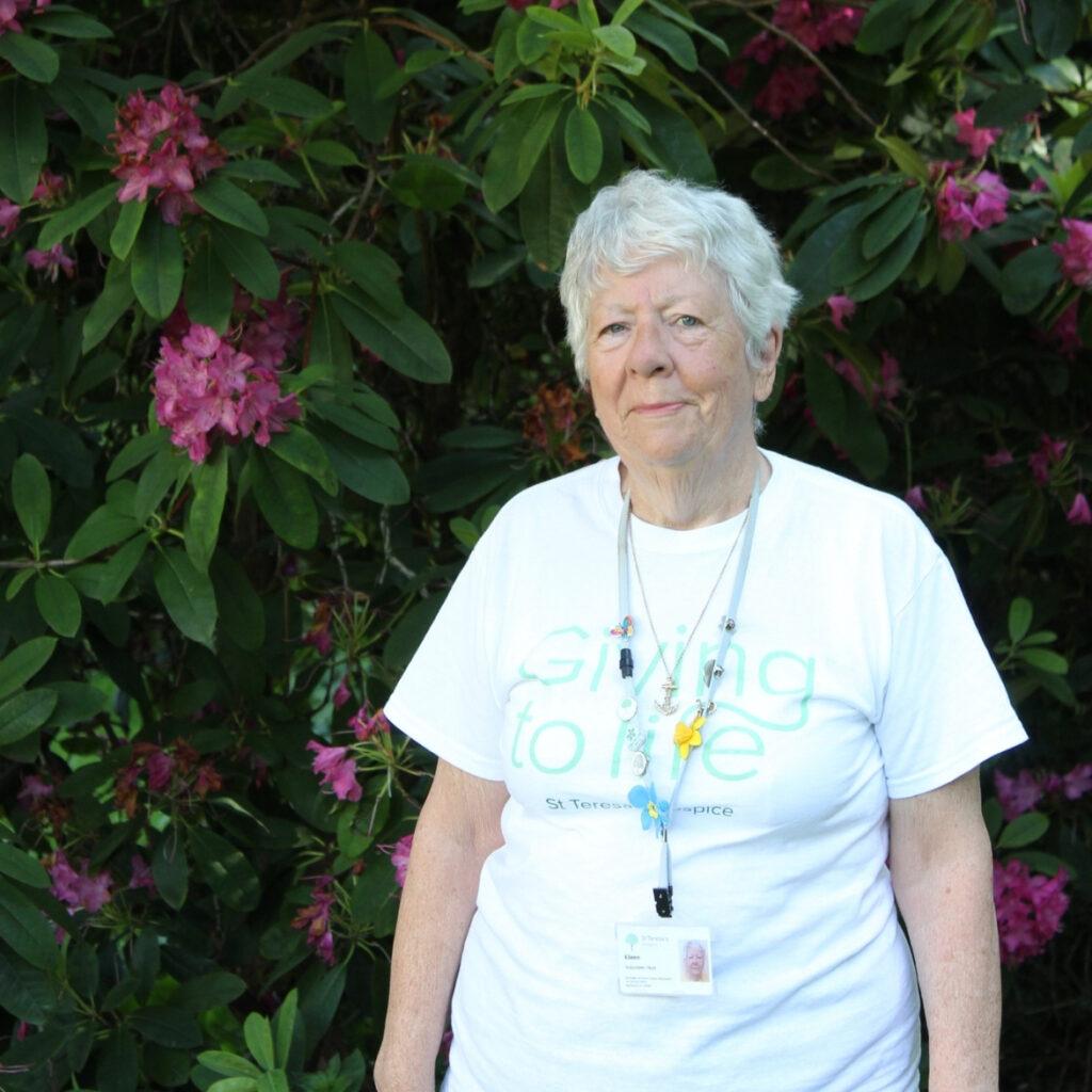 Lady in her mid-seventies standing in hospice gardens in front of lush dark green and pink foliage.